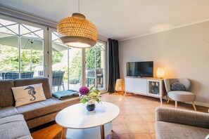 Ein modernes Wohnzimmer mit Sofa, Sessel, Fernseher und einem Tisch mit Blumen.