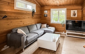 L’intérieur d’un chalet en bois, avec un canapé gris, une table basse blanche, un téléviseur et deux fenêtres ornées de plantes.