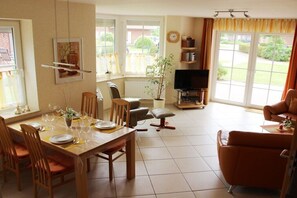 A dining area with a table set for four, a television, and a sofa in a bright room with large windows. - Holiday home with 2 bedrooms (Norddeich)