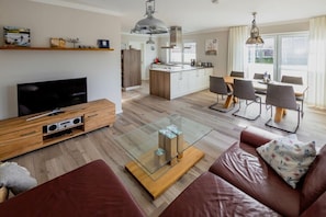 A modern living room with a TV, glass coffee table, leather sofa, dining area, kitchen, and large windows.