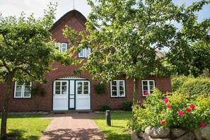 A brick house with a white door and windows, surrounded by a garden with trees and bushes. - Holiday apartment in Nebel with swimming pool (Nebel)