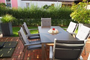 Eine Terrasse mit Tisch und Stühlen, auf dem Tisch ein kleiner Kuchen und ein Boden aus Ziegelsteinen.