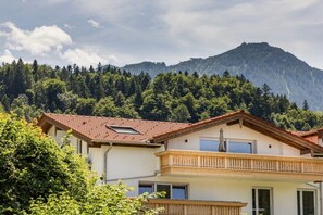 Ein zweistöckiges Gebäude mit Balkon, umgeben von Grün und Bergen im Hintergrund.