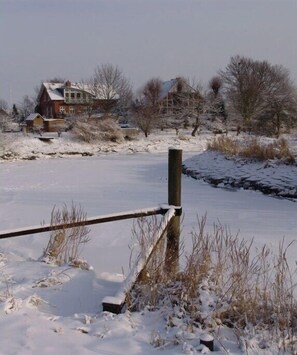 Schneebedeckte Landschaft mit Holzzaun, einem Haus im Hintergrund und einer zugefrorenen Wasserfläche.