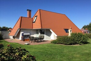 Ein Haus mit geneigtem Dach, Schornstein, einer Terrasse mit Tisch und Stühlen sowie einem gepflegten Rasen.