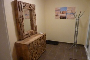 Ein Holzkabinent mit eingekerbten Lettern, ein Spiegel und eine Stehlampe in einem Raum mit Fliesenboden und einer Wand mit abstrakter Kunst.