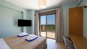 A hotel room with a bed, a TV, a balcony with curtains, and a wooden panel on the wall. - Alekos Beach Houses-Meliti Villa (Kefalonia)