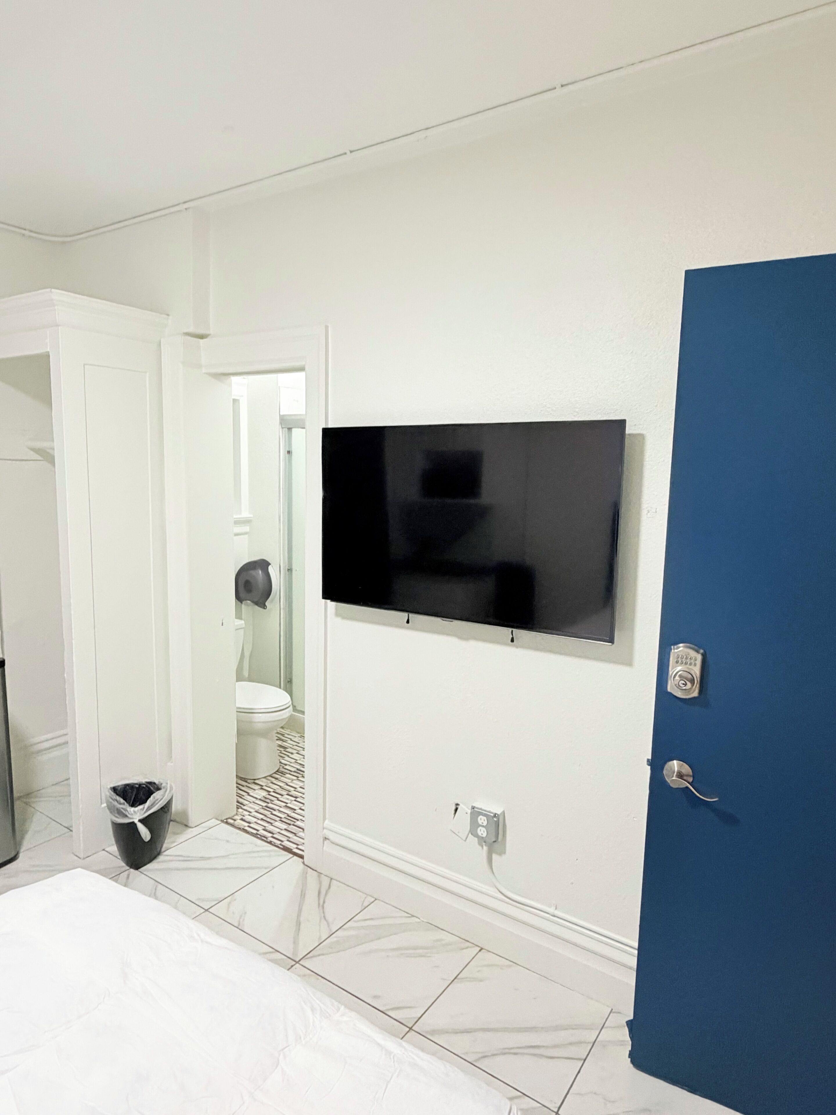 Quarto com cama, televisão fixada na parede, porta azul e sanita branca visível através de uma porta aberta.