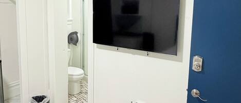 Quarto com cama, televisão fixada na parede, porta azul e sanita branca visível através de uma porta aberta.