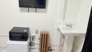 Een badkamer met een flatscreen televisie aan de muur, een magnetron op een klein wit apparaat, een radiator en een wastafel met spiegel.