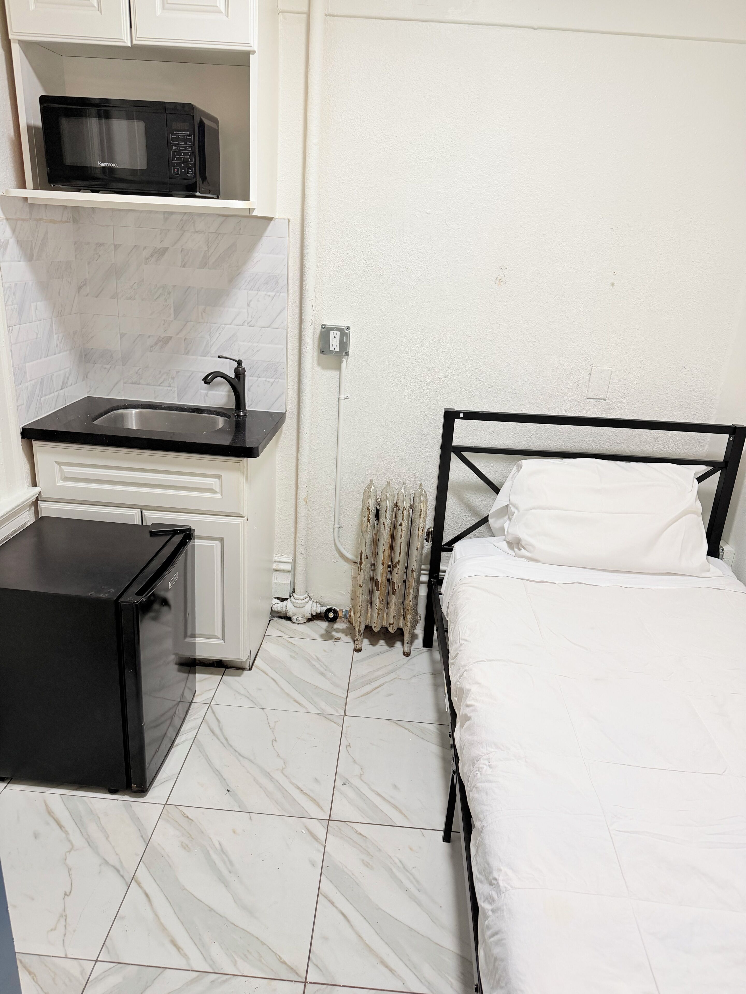 Una habitación compacta con cama, una kitchenette pequeña con microondas, un lavamanos y un radiador.