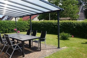 Eine Terrassengruppe mit Tisch und Stühlen, ein überdachter Pergola und ein Hecken im Hintergrund.