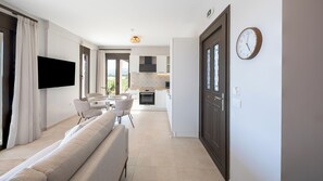 A modern living room with a sofa, a dining table, and chairs, a kitchen with an oven and cabinets, and a large window. - Alekos Beach Houses-Aelia Villa (Kefalonia)