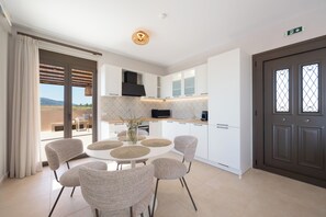 A modern kitchen with a round dining table, white cabinets, and a view through the glass door. - Alekos Beach Houses-Aelia Villa (Kefalonia)