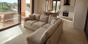 A living room with a beige sectional sofa, a dining table with chairs, a kitchen area, and a view of a swimming pool and greenery outside. - Alekos Beach Houses-Nereids Villas (Kefalonia)