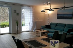 Ein modernes Wohnzimmer mit Sofa, Couchtisch und einem Esstisch mit Stühlen. Es gibt ein großes Fenster mit Vorhängen, eine Stehlampe und ein Bild an der Wand.