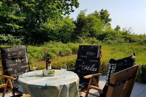 Ein Tisch im Freien mit Flasche und Glas, umgeben von Stühlen mit gemusterten Kissen, vor einer grünen Kulisse.