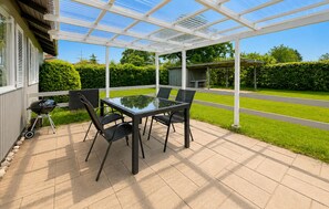 Eine überdachte Terrasse mit einem Glastisch und Stühlen, einem Grill und Blick auf eine grüne Rasenfläche und Bäume.