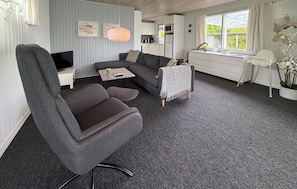Ein modernes Wohnzimmer mit einem grauen Sofa, einem Fernseher, einem kleinen Tisch mit einer Pflanze und einem offenen Durchgang zum Küchenbereich.