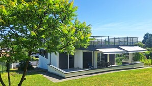 Una casa moderna con un porche cubierto, un árbol grande en primer plano y un cielo azul despejado.