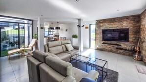 Una sala moderna con un sofá seccional, una mesa de centro de vidrio, un televisor de pantalla plana montado en una pared de piedra y un área de comedor al fondo.