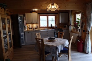 Ein Holzblockhaus-Innenraum mit Esstisch, Stühlen, einem Kühlschrank, Schränken, einem Fenster und einer Pendelleuchte.