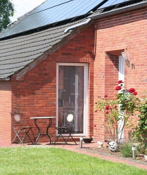 Un bâtiment en briques avec un toit en pente, une porte d’entrée en verre, une petite table et des chaises à l’extérieur, et un pot de fleurs avec des fleurs rouges.
