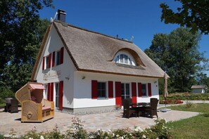 Ein traditionelles Haus mit Strohdach, roten Fensterläden und einem kleinen Garten mit Gartenmöbeln.