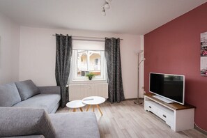 Ein Wohnzimmer mit einer Couch, einem Fernseher auf einem Holzboden, einem kleinen Tisch und einem Fenster mit Vorhängen.