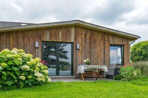 A wooden building with a large glass door, a small table with chairs, and a garden with hydrangeas. - Holiday apartment "Karla" (Ahrenviölfeld, Deutschland)