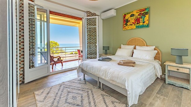 Un dormitorio con cama, mesita de noche, lámpara, cuadro y vista al océano.
