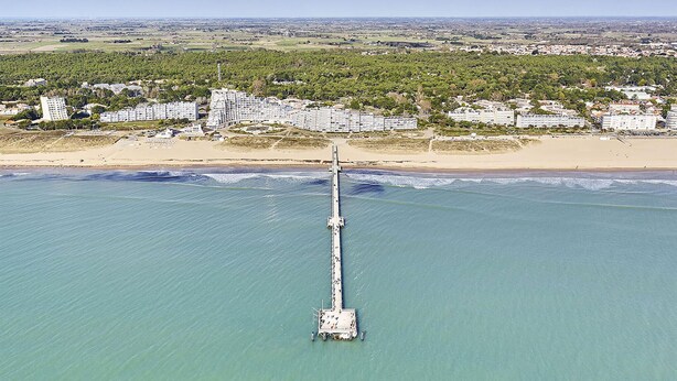 Une vue aérienne d’une zone côtière avec une longue jetée qui s’avance dans la mer, entourée de bâtiments et de verdure.