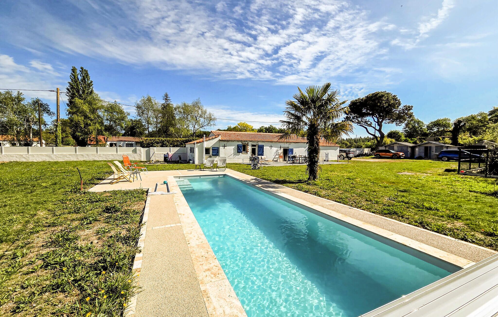 Una piscina rectangular con superficie de agua azul transparente, rodeada de césped, algunos árboles y un edificio pequeño al fondo.