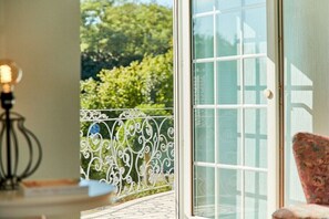 Ein Zimmer mit einer Schiebetür, die auf einen Balkon mit schmiedeeisernem Geländer und Blick auf Bäume führt.