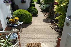 Ein schmaler, gepflasterter Weg führt zu einer Tür, gesäumt von Blumentöpfen und einem kleinen Tisch mit gelben Blumen.