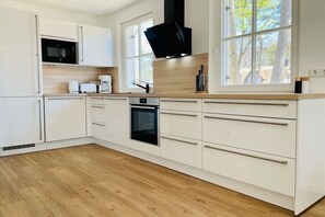 A modern kitchen with white cabinetry, a built-in oven, a microwave, and a large window with a view of trees. - Holiday home Lake & Countryside (Zirchow)