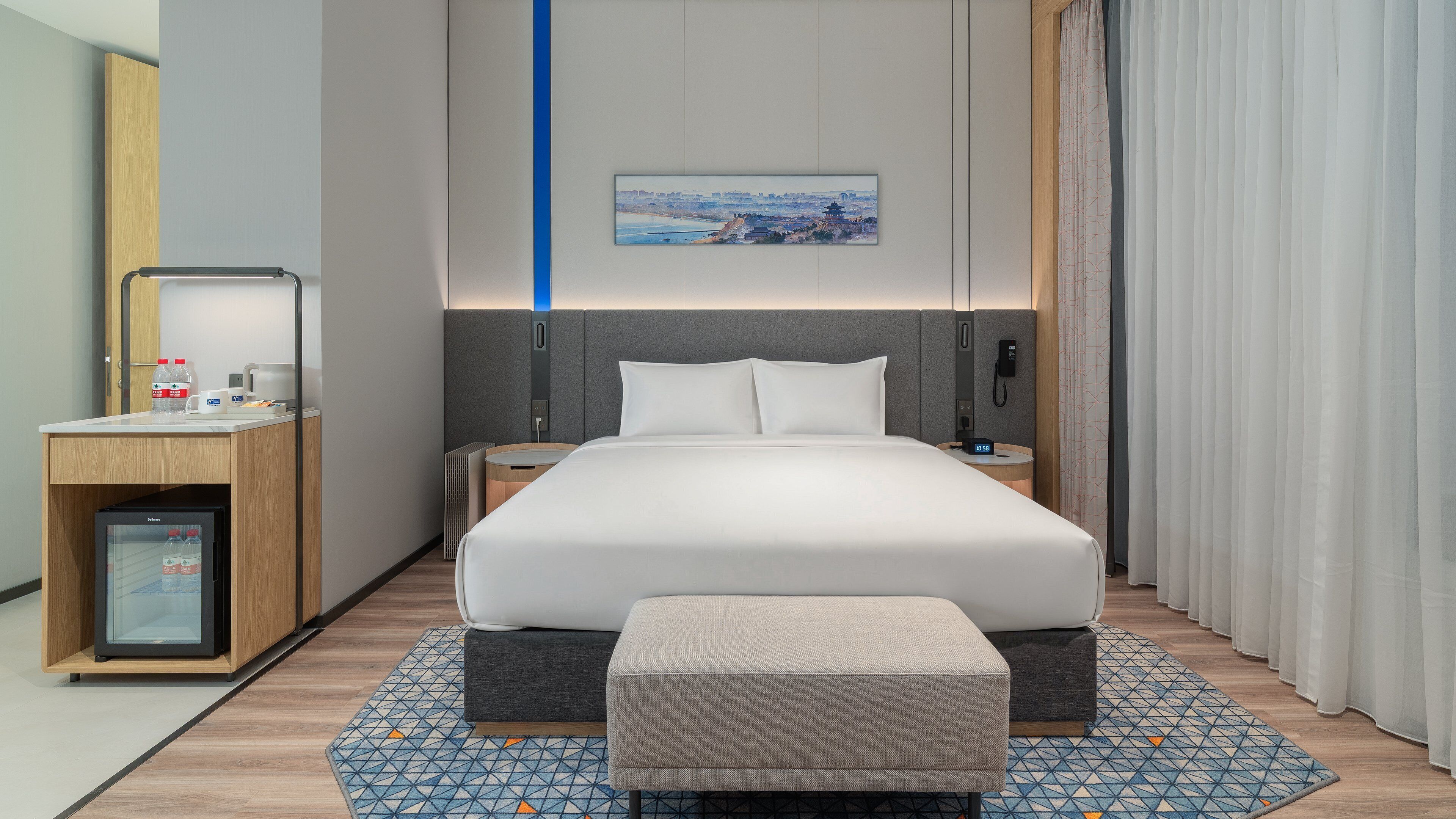 A modern hotel room with a large bed, bedside tables, a small refrigerator, a bedside lamp, and a view of a cityscape.