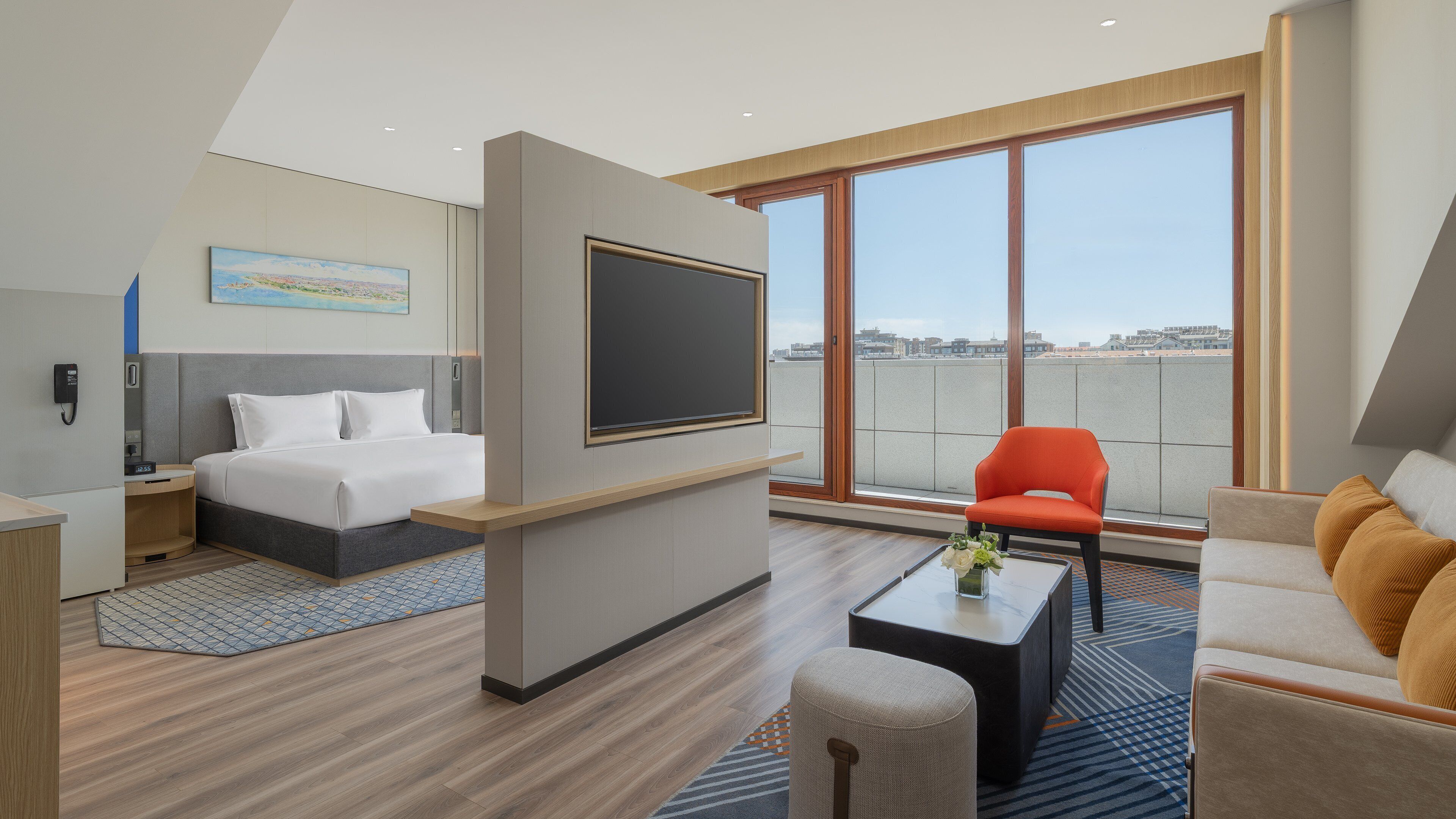 A modern hotel room with a bed, a flat-screen TV, a sofa, a coffee table, and a large window offering a city view.