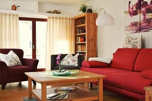 Ein Wohnzimmer mit einem roten Sofa, zwei Sesseln, einem Holztisch, einem Bücherregal und einem Bild an der Wand.