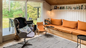 Quarto aconchegante com teto de madeira, um sofá com almofadas laranja, uma poltrona, uma mesa pequena e uma estante com livros.