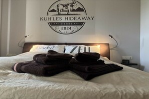 Ein ordentlich bezogenes Bett mit gefalteten Handtüchern, ein an der Wand befestigtes Schild mit einer Kuh-Illustration und Schriftzug, sowie eine Nachttischlampe.