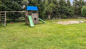 Una estructura de juego de madera con tobogán y columpios, rodeada de un césped y árboles.
