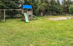 Eine hölzerne Spielanlage mit Rutsche und Schaukeln, umgeben von einem Rasen und Bäumen.