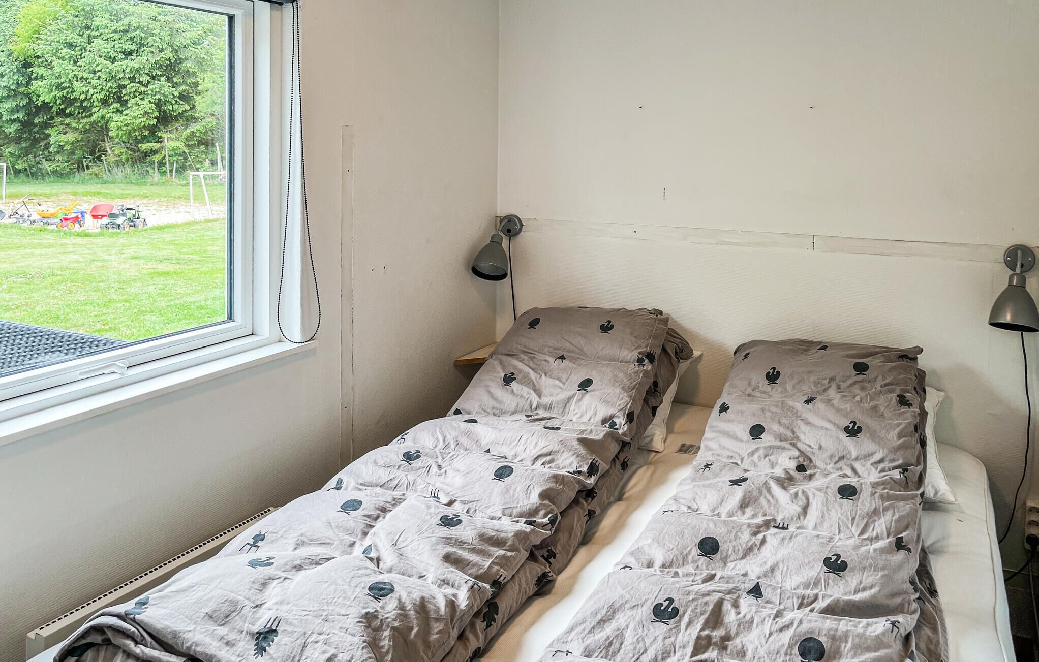 A small, single-bed room with a window, two bedside lamps, and a view of a garden.