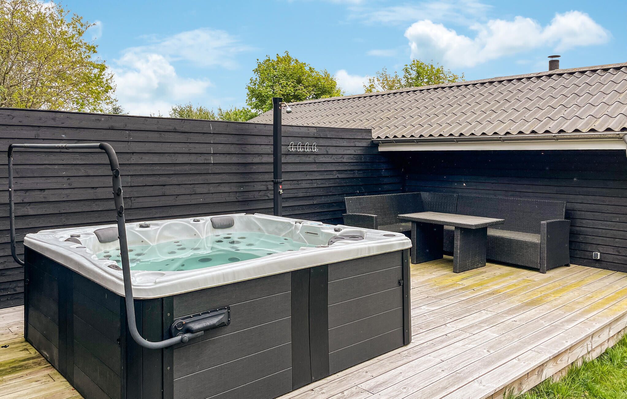 Un jacuzzi en una terraza de madera, con una mesa y sillas de patio, rodeado por una valla de madera y un edificio con tejado de baldosas.