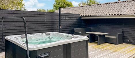 Un jacuzzi en una terraza de madera, con una mesa y sillas de patio, rodeado por una valla de madera y un edificio con tejado de baldosas.
