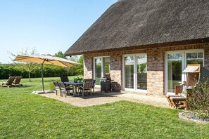 Une maison en briques avec un toit de chaume, une terrasse avec une table et des chaises, et un barbecue.