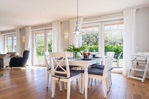 Een eetgedeelte met een witte tafel, witte stoelen, een hanglamp en grote ramen met witte gordijnen.