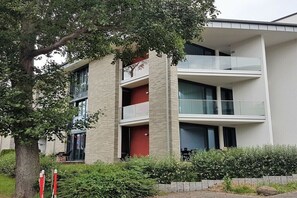Ein modernes mehrstöckiges Gebäude mit Balkonen, ein Baum im Vordergrund und ein Hecken im Hintergrund.