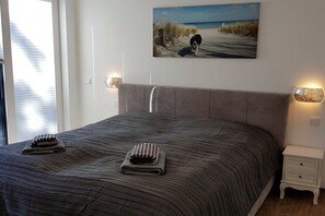 Ein Bett mit gestreifter Bettdecke, zwei Handtücher, ein Gemälde mit Strandmotiv und ein Nachttisch mit Lampe.